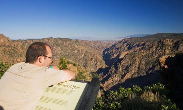 A person leans over an interpretive sign and gazes into the distance at the striped walls of the Black Canyon of the Gunnison National Park near Montrose, Colorado