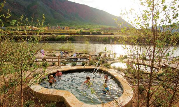 A group of people soaks in a stone hot pool overlooking a river and the mountains