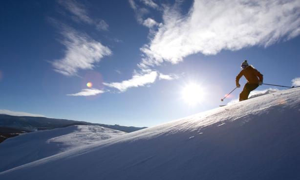 A skier shoots down a slope under a blue sky with a bright sun lighting up the scene.