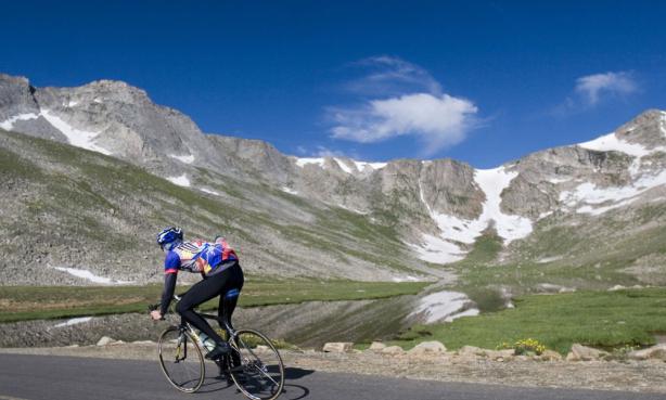 A cyclist zooms up a roadway under bright blue skies and a grassy meadow and a mountian ridge with a trace of snow
