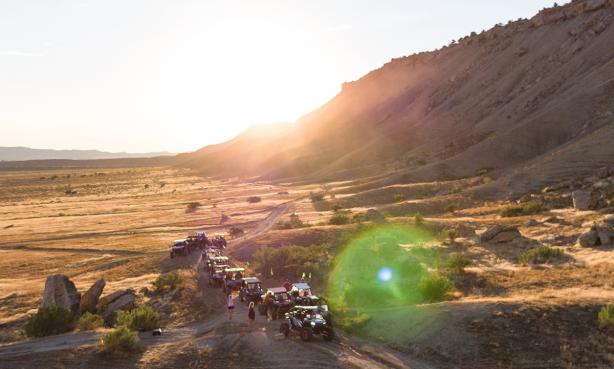 A group of off-road vehicles in a line on a dusty trail on the mesa. The sun is peeking through the plateau hill tops behind them, shining on the vast, dusty flatlands.