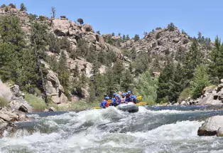 A group of people dressed in blue safety equipment and holding yellow paddles sit on an inflatable grey raft as it flows down the Arkansas River near Buena Vista. The whitewater waves are mild. Behind the whitewater rafters are hills of craggy rock formations sprinkled with pine trees, all under a clear blue sky.