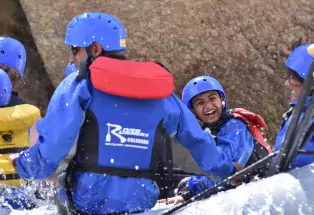 A photograph of a kid smiling widely, clad in a blue helmet, blue jacket and a life vest. They are on a whitewater rafting trip with their family on the Arkansas River in Colorado. Beside the kid are several smiling adults. There is water splashing up as they raft down the river.