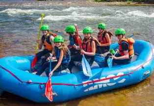 A group of six smiling middle-school-aged kids with neon-green helmets and orange safety vests sit on a large blue inflatable raft. They are all holding a raft as they prepare to be pushed into the calm waters of the Arkansas River behind them.