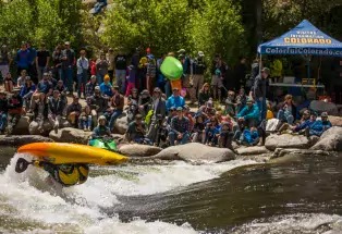 A professional kayaker in a yellow-and-orange kayak is fully flipped upside down as they fly above the whitewater waves of the Arkansas River at the Buena Vista CKS Paddlefest. Behind them is a group of spectators along the side of the river, watching intensely. In the corner is a tall blue tent that says "Visitor information Colorado," and "ColorfulColorado.com." The sun is shining on everyone at the festival.