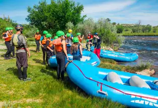 A group of Arkansas River whitewater rafters stand by their inflatable blue rafts, clad in bright green helmets and orange vests. Guides are holding lines and demonstrating how to safety raft on the river, near Buena Vista. The river is rolling in front of them past a little green pasture and the sun is shining down.
