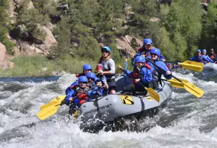 A group of smiling and focused people dressed in blue safety equipment and holding yellow paddles rushes down the Arkansas River on a whitewater rafting trip. They are actively rafting on a gray inflatable raft at the top of the Zoom Flume rapid in Browns Canyon National Monument.