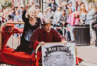Madam Lou Bunch Bed Race with man in hat holding onto footboard, woman dressed in black behind him on the bed and man in red pushing the bed down the Main Street. 