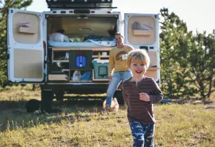 Parent watches child play in the grass from the open back of a campervan. 