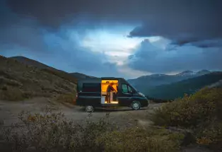 Person inside of a lit campervan with the side door open as the sun sets behind the mountains.