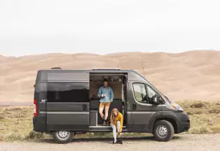 Two people sit in the open side doorway of a campervan with the dunes of Great Sand Dunes National Park in the background.