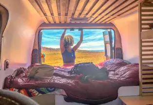 Woman closes door on the back of a campervan with cozy bedding and there are prairie grasses and blue skies in the background