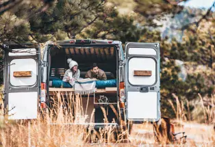 Two people wearing winter clothes lay on their stomachs in a campervan bed and look out the open back door.