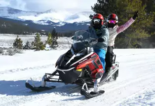 Two people wearing helmets, goggles and winter gear ride a red snowmobile near Leadville with snow-covered mountains in the distance.