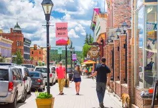 People stroll along the shop-lined sidewalk in downtown Leadville on a sunny summer day with cars parked to the left and a bright blue sky up above.