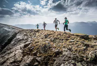 Four runners jog along a rocky ridge near Leadville with mountains and clouds against a blue sky in the background.