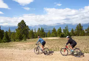 Two cyclists enjoy mountain biking along a gravel trail near Leadville with evergreen trees, a bright-blue sky and fluffy white clouds in the distance.
