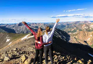 Two women at the top of a hike facing mountains with a blue sky above.