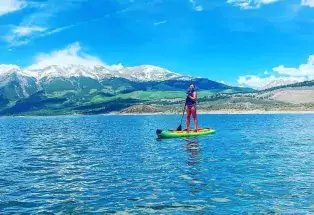 A woman stands on a stand-up paddleboard on a glittering lake with mountains in the background near Leadville.