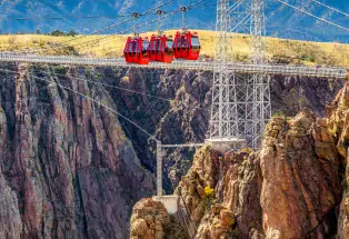 Royal Gorge Bridge & Park cherry-red aerial gondola crossing the gorge with two suspension bridge metal towers in the background. Mountain peaks are in the far background and rock outcropping are in the foreground.
