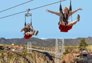 Two people sitting in red-bottomed chairs on the Zip Line. First person is a man yelling with legs out, second person is a girl with her arms out looking at the camera.