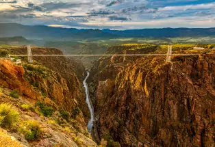 Royal Gorge Bridge & Park: Golden hour with Arkansas River snaking through rock walls of the gorge with the metal suspension bridge across the upper part of the canyon.