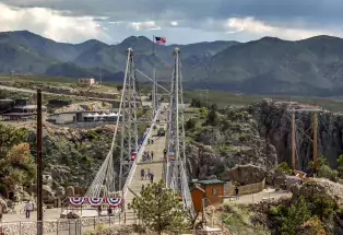 Royal Gorge Bridge with metal suspension towers in the foreground and mountain range in the background. People crossing the bridge and patriotic bunting. 