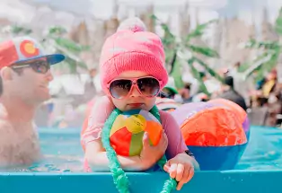 The coolest kid ever hanging out in a pool with sunglasses and a pink cap.