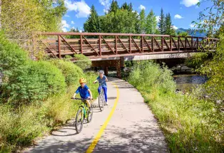 Kids biking near a river with a bridge in the background.