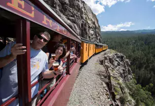 People holding glasses of beer pose in Durango Train windows as the train travels on a cliffside