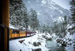 The orange Durango Train starkly contrasts with the bright white snow on the ground and dusting trees as well as the teal of the river running alongside the train