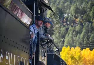 Two train engineers in historical 1800s dress lean out slightly from the engine in front of a golden stand of aspens
