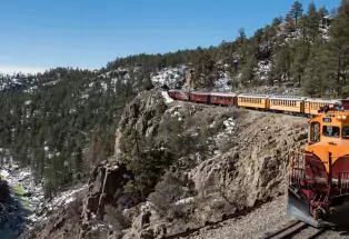 The Durango Train travels alongside a cliff with patches of snow on the ground