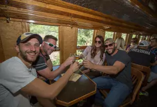 A group of four people clink beers as they sit on a historical train car with polished wooden walls and ornate carvings
