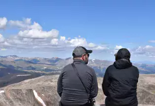 Two people sit at the 14,265-foot summit of Mount Blue Sky looking at other mountains in the distance