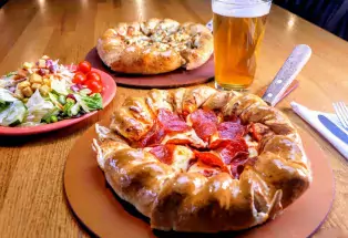 A pepperoni pizza with a very thick crust pictured with a beer a salad and another pizza in the background