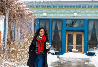 A person holding a to-go cup of tea walks out of Boulder Dushanbe Teahouse's peacock blue and teal tiled doorway on a walkway lined with snow
