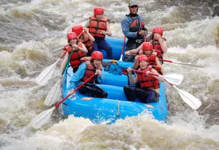 Eight people wearing helmets paddle in a blue raft through whitewater on the Colorado River