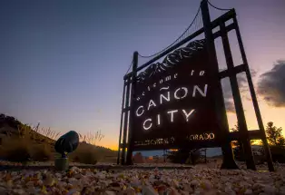 A tall metal sign with a replica of the Royal Gorge that says "Welcome to Cañon City." In the background is a deep purple and yellow sunset with minimal clouds.
