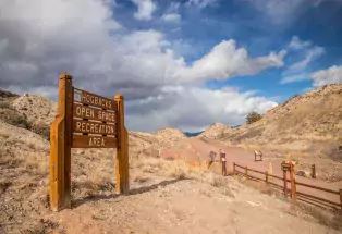 A wooden sign in the foreground labeling a trailhead that says "Hogbacks Open Space Recreation Area." In the background is a cloudy sky above a hiking trail path.