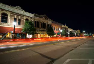 A strip of storefronts in downtown Cañon City at night. Red lights from cars in motion make a few steady streaks above the road.