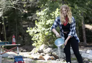 A camper fully douses her campfire with water from a silver bucket in the morning sunlight