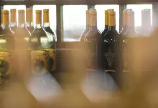 Wine bottles sit on a shelf at the Winery at Holy Cross Abbey behind a slightly blurry object. Behind the bottles are windows letting in natural light.