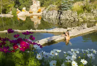 A woman rests on the edge of the Strawberry Park Hot Springs pool on a sunny day, with pink and white wildflowers blooming in the foreground
