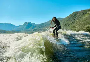 A woman in a black wetsuit surfs a wave on Grand Lake atop a wakeboard, with blue sky and green mountains behind her