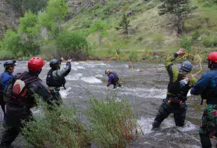 Six students in protective gear and backpacks stand knee-deep in whitewater waves of a river in Colorado. One of them is in the middle of the river while the rest are more on the side of it. Between them, there is a thin yellow rope frozen in the air, being tossed between them as part of the course. In the background is a tall, steep, green hill with scattered small trees and rocks.