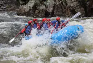 A group of whitewater rafters in red protective gear smiling and laughing on a blue raft. The raft is being tossed by the rushing water, splashing the people's faces. They all have paddles in their hands. Behind them on the raft is the instructor, barely visible, with their paddle in the water. In the background are rocky cliff-like structures beside the water.