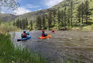 A scenic photo of calm river water running between two grassy areas. There are three student kayakers in the water on blue and orange kayaks wearing protective gear. They are paddling in the water facing away from the camera. In the background are grassy and rocky hills spotted with tall pine trees beneath a mostly blue sky.