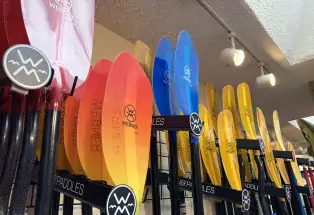 A close-up image of red, orange, yellow and blue water sports paddles hung upright on hooks against a beige wall. The paddles are labeled, "WERNER" and are staggered along the wall. Above them are popcorn ceilings and two small spotlights.
