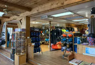A photo of a retail store adorned in wood paneling on the floors and ceiling. There are a variety of items on shelves, hooks and turntables, including sunglasses, balaclavas, hats, t-shirts, shoes, maps, kayaks, vests, backpacks and more.